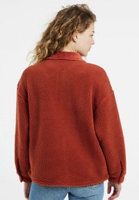 Fleece jacket in burnt orange with a textured, fluffy surface. Features a corduroy collar and oversized fit. Long sleeves with elastic cuffs.