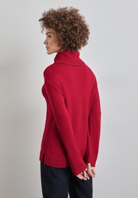 Roter Rollkragenpullover mit geripptem Muster, seitlichen Schlitzen und langen Ärmeln. Glattes Material, lockerer Schnitt und kontrastierendes Kragendetail.
