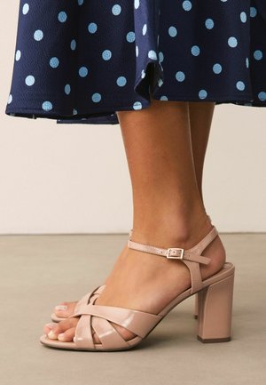 Gebräunte Beine, die nudefarbene Sandalen mit Blockabsatz aus lackiertem Leder und Knöchelriemen tragen, kombiniert mit einem marineblauen Rock mit hellblauen Polka-Dots.