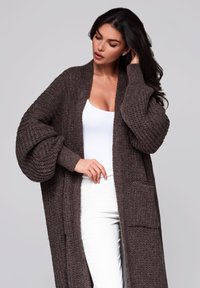 Ruskea neulottu cardigan, jossa on teksturoitu kuvio, ylisuuria hihoja, avoin etuosa ja sivutaskut. Valittuna valkoisen paidan ja vaaleiden housujen päälle.