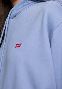 Person som bär en ljusblå huvtröja med en röd Levi’s-logotyp broderad på bröstet, syns del av långt mörkt hår.