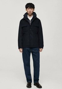 Svart ullblandad jacka med dragkedja på huvan, två stora bröstfickor och knappstängning; tillsammans med mörkblå jeans och svarta skor.