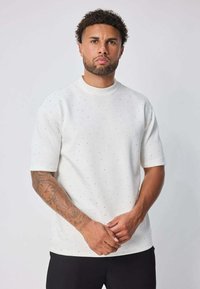 Chemise blanche à manches courtes avec de petits pois noirs, col rond et un tissu légèrement texturé. Le modèle se tient avec les bras croisés.