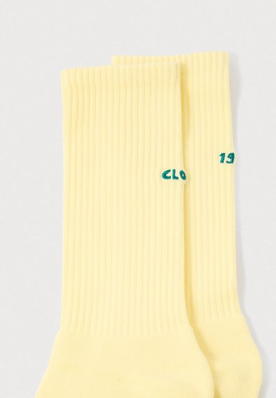 Κίτρινες ριγωτές κάλτσες με πράσινο κεντητό κείμενο "CLO" και "19" στην κορυφή. Μαλακό βαμβακερό υλικό με στάνταρ σχεδιασμό μήκους έως τον μέσο της γάμπας.
