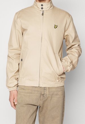 Veste beige zippée avec poignets et ourlet élastiques, col à boutons, et petit logo d'oiseau jaune sur la poitrine gauche, portée avec un pantalon marron clair.