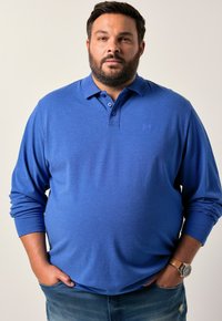 Sininen pitkähihainen polo-paita, joka on valmistettu pehmeästä kankaasta. Paitassa on kolmen napin lista ja huomaamaton logo rinnassa. Yhdistetty farkkuihin.