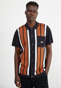 Camisa de botones de manga corta con rayas verticales en negro, marrón y blanco. Cuenta con un bolsillo en el pecho con un parche de logo. Tejido de algodón.