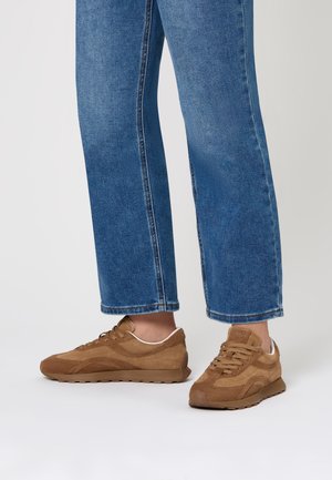 Scarpe in suede marrone con profilatura bassa, tomaia testurizzata e dettagli bianchi. Presentano lacci rotondi e suola in gomma con battistrada. Sono visibili pantaloni di jeans.