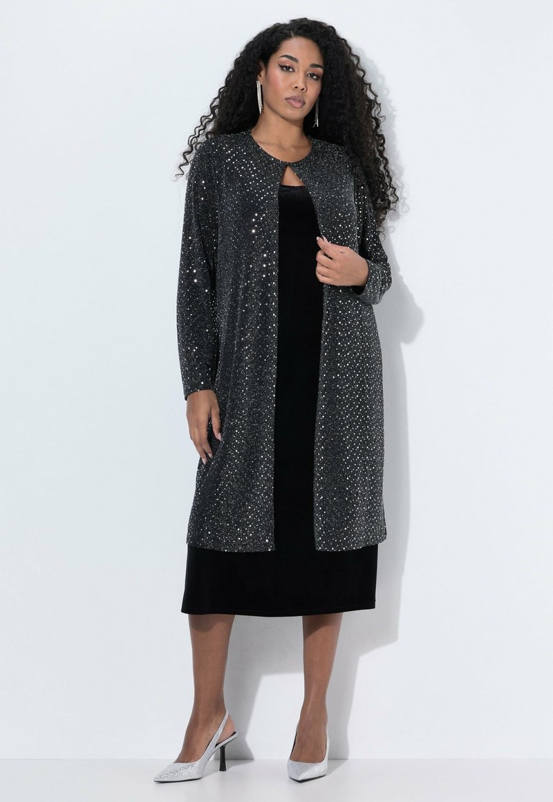 Vestido de terciopelo negro con un cárdigan largo, abierto por delante, cubierto de lentejuelas plateadas. Cuello alto y mangas tres cuartos. Tacones plateados completan el conjunto.