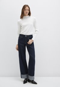 Weißer, gestrickter Pullover mit hohem Kragen und strukturiertem Muster, kombiniert mit dunkelblauen Jeans mit weitem Bein und schwarzen Stiefeln mit Absatz.