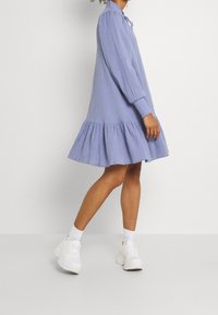 Vaalean sininen pitkähihaine mekko, jossa on poimutettu helma, teksturoitu kangas ja solmimisella kiinnitys kaulassa. Yhdistetty valkoisiin lenkkarit ja sukkiin.