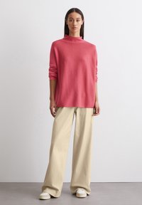Roze trui met hoge hals, losse pasvorm en geribde manchetten, gecombineerd met wijde beige broek en witte sneakers op een neutrale achtergrond.