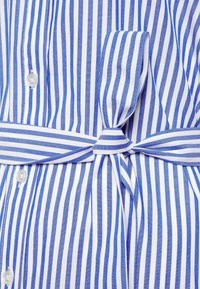 Camisa a rayas azul y blanca con un lazo en la cintura, que presenta un frente de botones y una textura de algodón suave. Las rayas varían en ancho.