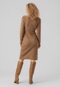 Robe en tricot de couleur marron chaud avec des manches longues et une silhouette ajustée, présentant une texture côtelée. Associée à des bottes à talons beiges jusqu'au genou.