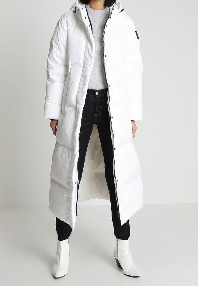 Lunga giacca puffa bianca con cappuccio, chiusura frontale con zip, polsini elasticizzati e tasche laterali. Indossata sopra una maglietta grigia e jeans neri.