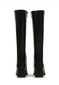 Bottes noires en cuir synthétique arrivant jusqu'aux genoux, avec un design ajusté, des talons carrés et des zips à l'arrière. Texture lisse et minimalisme dans les détails.