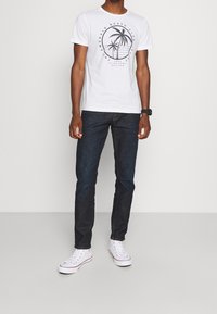 Camiseta gráfica blanca con diseño de palmera, jeans azul oscuro y zapatillas blancas con acentos rojos. Un atuendo casual sencillo.