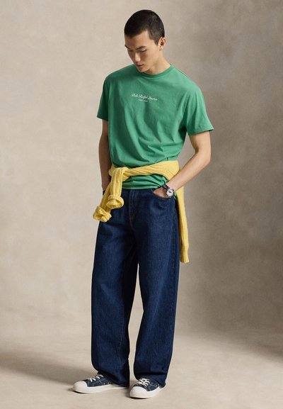 Νέος άνδρας που φοράει πράσινη μπλούζα Polo Ralph Lauren, φαρδιά μπλε τζιν, κίτρινο πουλόβερ δεμένο γύρω από τη μέση και ναυτικό μπλε αθλητικά παπούτσια.