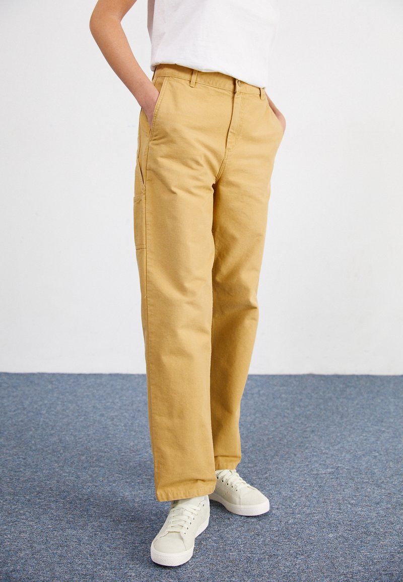 Pantalones de algodón beige con corte recto, dos bolsillos laterales y un bolsillo cargo, combinados con zapatillas verdes claras.