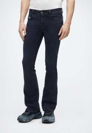 Mężczyzna w czarnych jeansach slim fit, czarnych trampkach i białej koszuli, stojący na jednolitym jasnym tle.