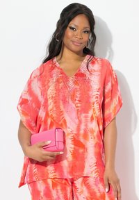 Blusa a tie-dye nei toni del rosa brillante e arancione, con scollatura a V e maniche corte, arricchita da decorazioni sul collo. Abbinata a una pochette rosa.