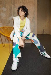 Mönstrade breda byxor i flera färger med geometriska former, kombinerade med en gul topp och en vit zip-up hoodie, båda bärs med vita sneakers.