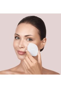 Applicateur facial en silicone à la forme irrégulière, de couleur blanche, tenu contre la joue d'une personne à la peau lisse et sur un fond neutre.