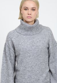 Grå strikket sweater med høj, foldet krave. Blød tekstur og afslappet pasform. Lange ærmer med en let oversized silhuet.