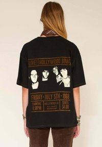 Personne portant un T-shirt de concert vintage noir affichant la liste des groupes live du Hollywood Bowl de 1968 et les détails des billets en impression marron.