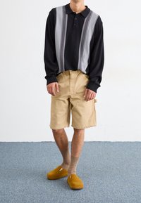 Schwarzes Langarm-Polo mit grauen vertikalen Streifen, kombiniert mit beigen Cargo-Shorts. Gelbe Clogs und braune Socken runden das Outfit ab.