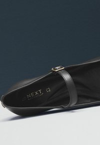 Černé kožené Mary Jane ploché boty s tenkým nastavitelným páskem a zlatou sponou, uvnitř označené NEXT MotionFlex.