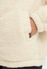 Kreemjas värvitoonis fliispulm, millel on karvane tekstuur, külgpocket ja väike pruun brändi silt ääres.