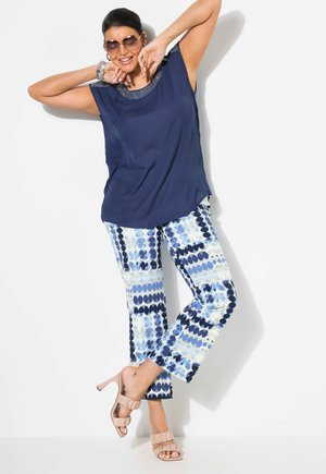 Marineblauw mouwloos topje met een vergulde halslijn, gecombineerd met gemusterde blauw-witte broek. Model draagt lichtroze hakken.