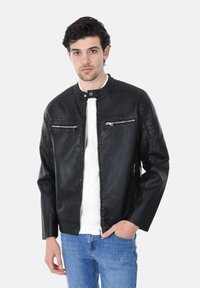 Giacca in pelle nera con un design aderente, colletto rotondo, tasche con zip e dettagli testurizzati sulle spalle. Indossata sopra una camicia bianca e jeans blu.
