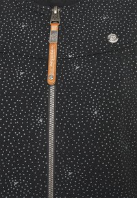 Svart zippered hoodie med ett prickmönster i grå nyanser, en läderdragrem och en metallogodetail nära axeln.