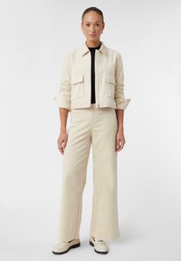 Krótkie beżowe żakiet z kieszeniami bocznymi i zapięciem na zamek, w połączeniu z szerokimi beżowymi spodniami oraz kremowymi butami z czarną podeszwą.