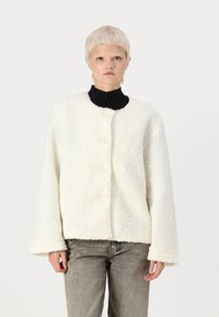 Veste en polaire couleur crème avec un col rond, fermeture à quatre boutons et manches larges. Surface texturée avec une apparence douce et chaleureuse.