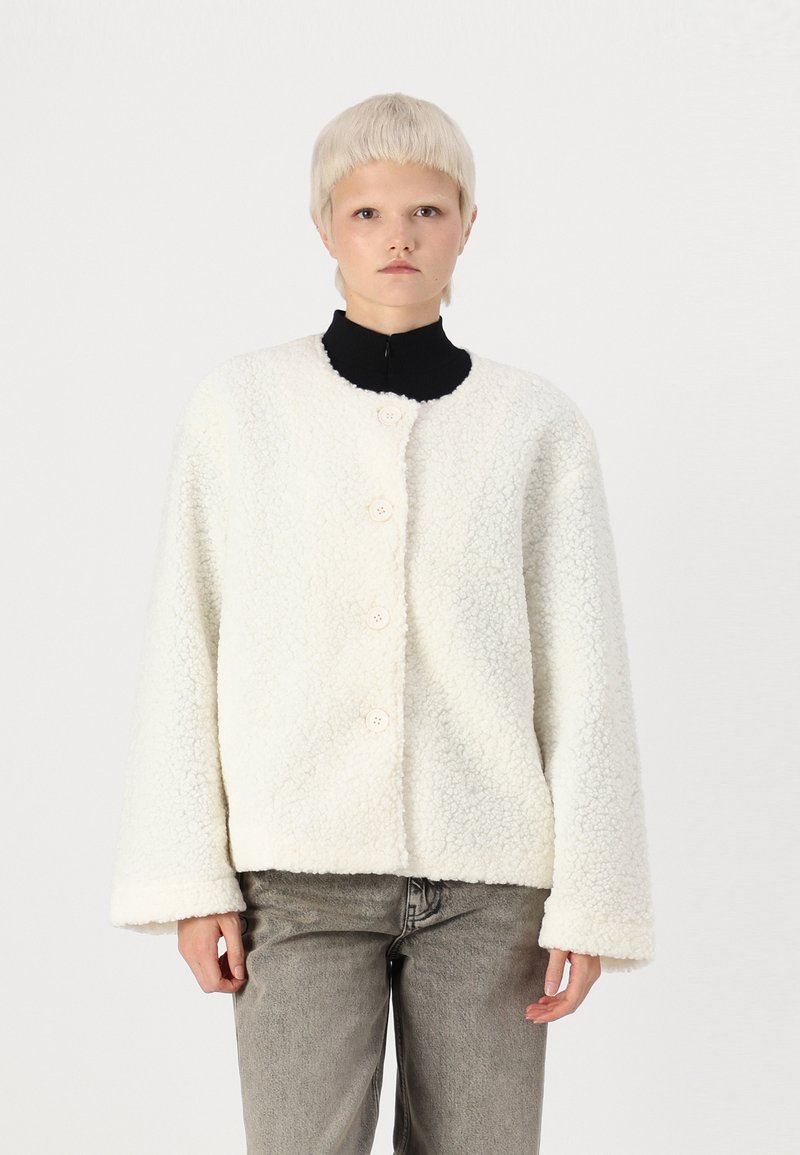 Veste en polaire couleur crème avec un col rond, fermeture à quatre boutons et manches larges. Surface texturée avec une apparence douce et chaleureuse.