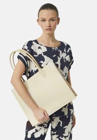 Beżowa skórzana torba typu tote z dwiema długimi rączkami, prostokątna, z subtelnym logo i minimalistycznym wzornictwem, trzymana przez modelkę.