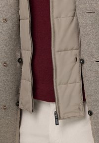 Cappotto esterno beige con uno strato di lana grigia testurizzata, aperto con cerniera per mostrare un maglione lavorato a maglia color borgogna e pantaloni bianchi in velluto a coste. Dettagli con bottoni distintivi.