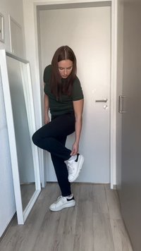 Białe sneakersy z czarnymi akcentami, noszone z ciemnymi spodniami. Osoba dostosowuje lewy but, stojąc obok lustra i drzwi.