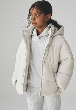 Veste matelassée beige clair avec capuche, dotée d'un rembourrage épais et d'une fermeture éclair. Superposée avec un sweat-shirt gris à côtes et un pantalon assorti.