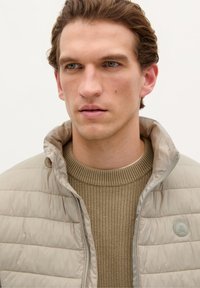 Beige puffer jacket with a textured knit green sweater underneath. Jacket features a high collar and subtle logo detail. Smooth, lightweight fabric.