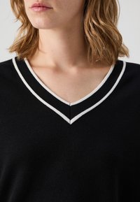 Pull en maille noire avec un col en V, orné d'une bordure rayée blanche le long de l'encolure. Texture douce et coupe décontractée.