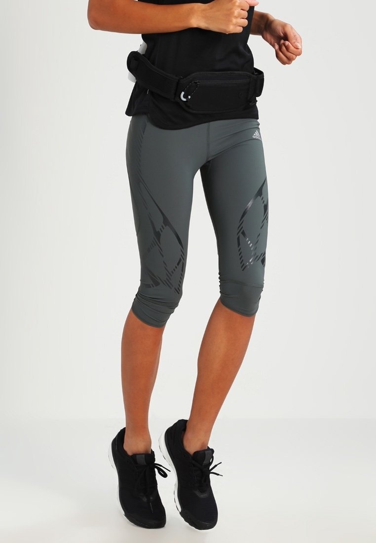 Szare, krótki legginsy z błyszczącym czarnym abstrakcyjnym wzorem oraz szerokim pasem w talii, w połączeniu z czarnym pasem fitnessowym i czarnymi butami sportowymi.