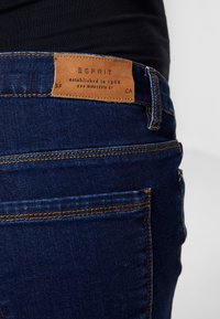 Mörkblå denimjeans med orange sömmar, med en läderetikett som visar varumärket "ESPRIT" och information.