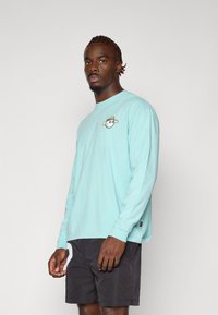 Turkis langermet t-skjorte med et tegneseriegraphikk på brystet, kombinert med mørke shorts. Mykt stoff og avslappet passform.