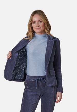 Marineblauer Cord-Blazer mit strukturierter Oberfläche, geöffnet, um ein gemustertes Innenfutter zu zeigen. Kombiniert mit einem hellblauen, glitzernden Rollkragenoberteil und passenden Hosen.
