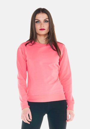 Femme aux longs cheveux bruns portant un haut corail rose à manches longues et un pantalon noir, debout devant un fond uni.