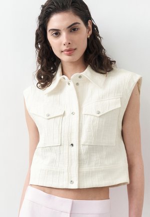 Femeie cu păr creț și închis la culoare, purtând un vestă albă fără mâneci, texturată, cu nasturi și buzunare cu clapetă, și pantaloni roz deschis.